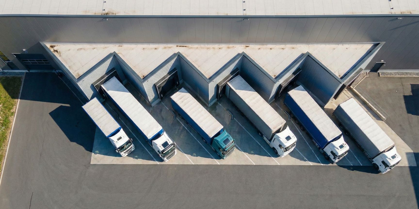 Luftaufnahme einer Logistikimmobilie mit Sägezahnrampen, an denen sechs Sattelzüge in Schrägaufstellung angedockt sind. Die Zickzack-Fassade der Verladehalle und die platzsparende Anordnung der LKW auf dem kompakten Vorfeld sind deutlich zu erkennen. Das Bild demonstriert die Schwalbenschwanz-Andienung in der Praxis.